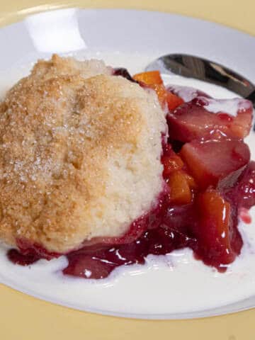 This photo for the Peach and Plum Cobbler shows the fruit in a bowl with a biscuit on top swimming in cream.