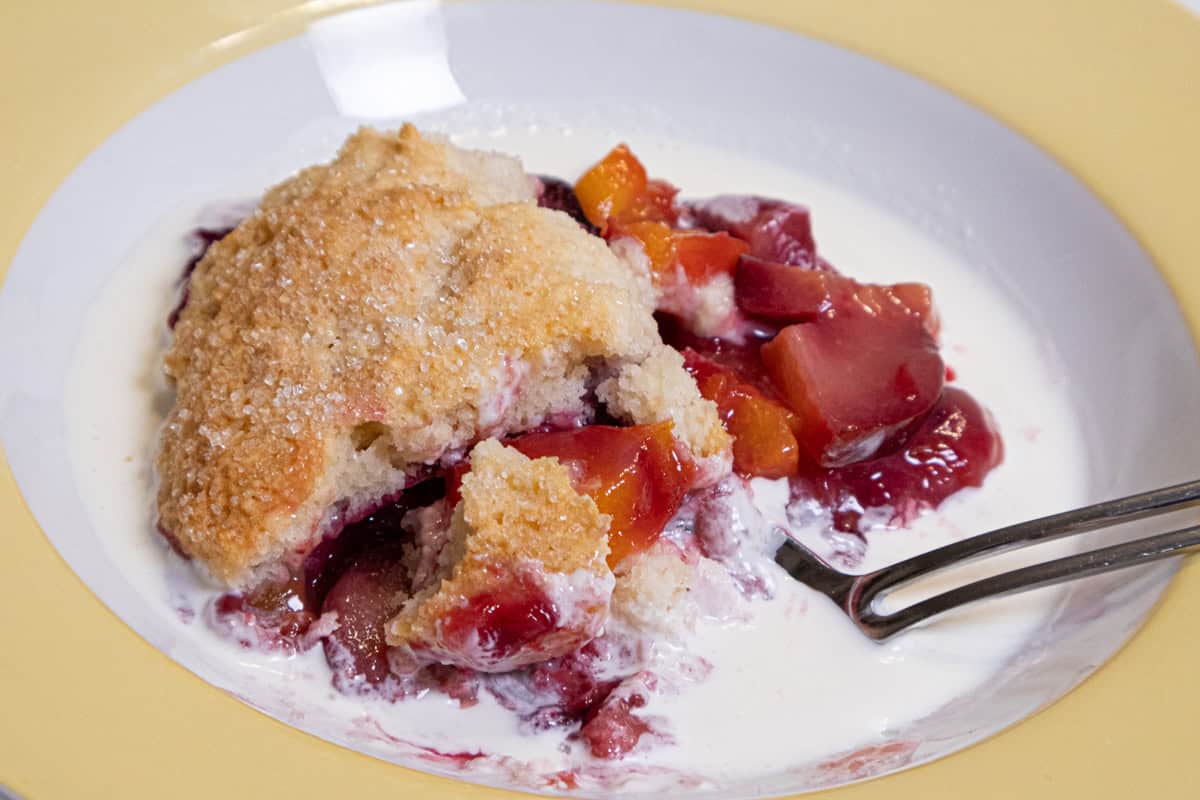 This photo of the Peach and Plum Cobbler shows the dessert in a yellow rimmed bowl swimming in half and half.