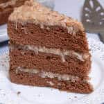 A slice of the Traditional German Chocolate Cake sits on a white lace edged plate with the whole cake behind it and a server on the side.