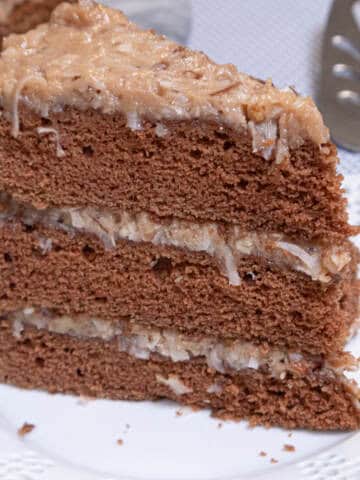 A slice of the Traditional German Chocolate Cake sits on a white lace edged plate with the whole cake behind it and a server on the side.