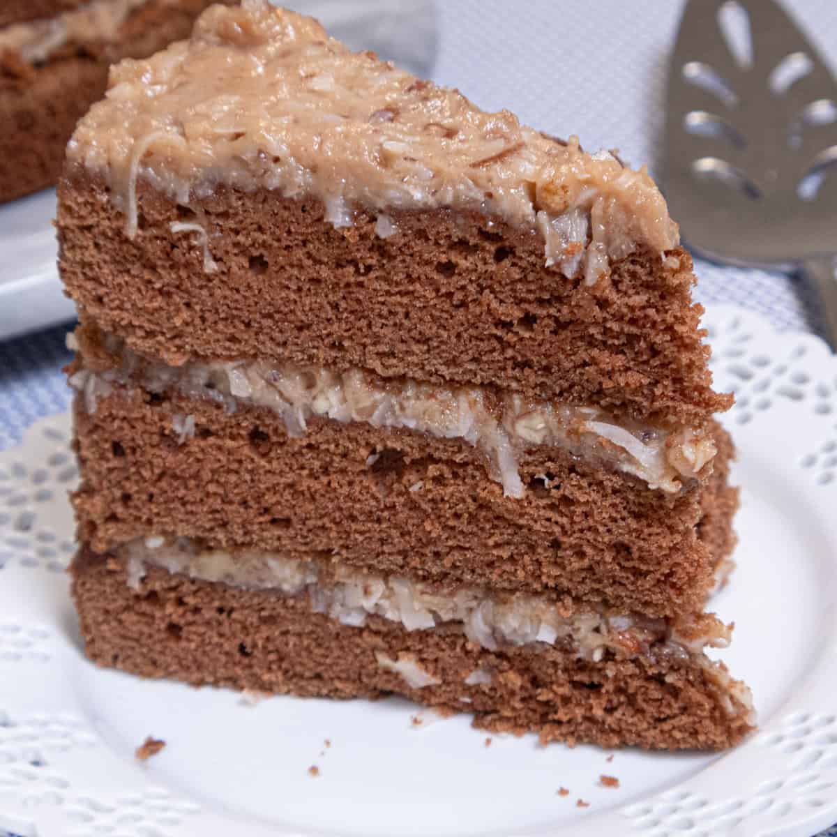 A slice of the Traditional German Chocolate Cake sits on a white lace edged plate with the whole cake behind it and a server on the side.