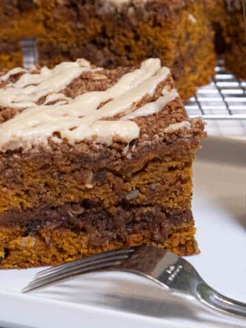 A piece of the Pumpkin Streusel Coffee Cake sists on a white plate with a fork and a cup next to it and more pieces on a cooling rack behind it.