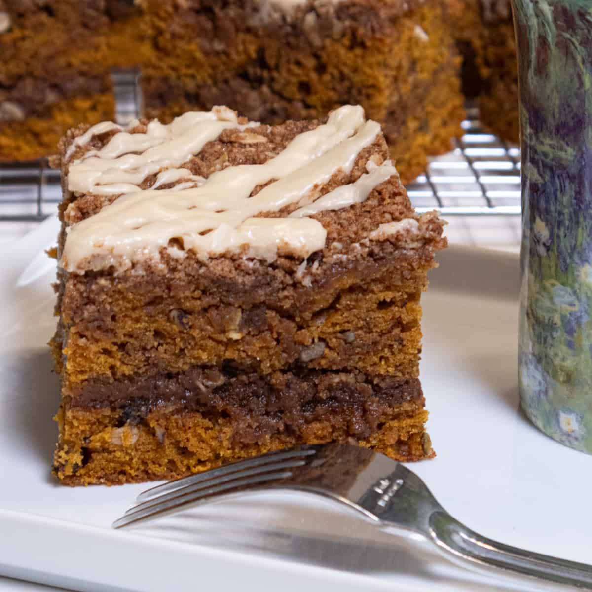 A piece of the Pumpkin Streusel Coffee Cake sists on a white plate with a fork and a cup next to it and more pieces on a cooling rack behind it.