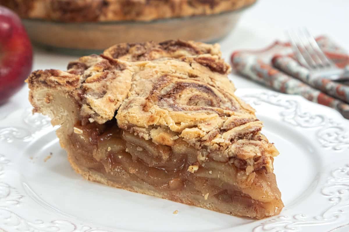 A slice of the Cinnamon Bun Apple Pie sits o a white plate with a napkin and fork next to it and the pie plate and apple in the background,
