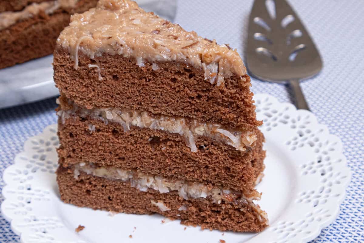 A slice of the Traditional German Chocolate Cake sits on a white lace edged plate with the whole cake behind it and a server on the side.