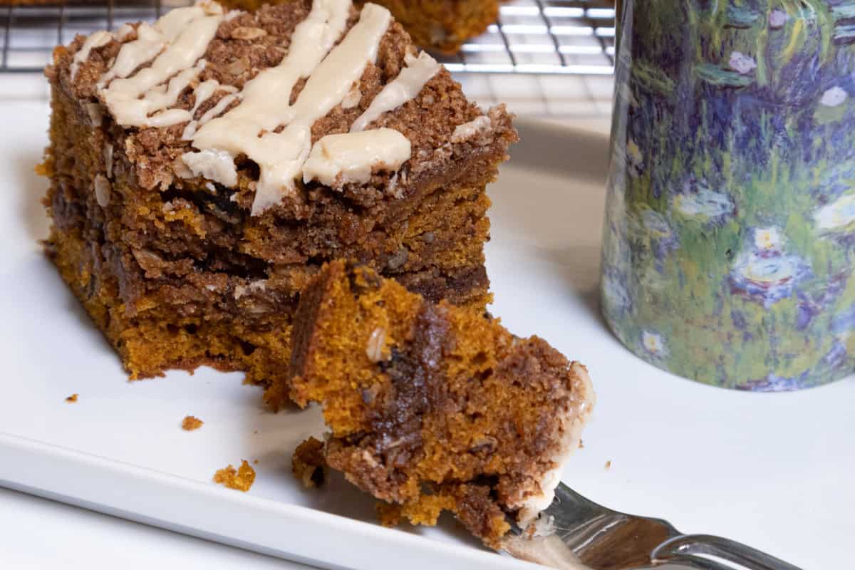 A piece of the Pumpkin Streusel Coffee Cake sists on a white plate with a fork and a cup next to it and more pieces on a cooling rack behind it.