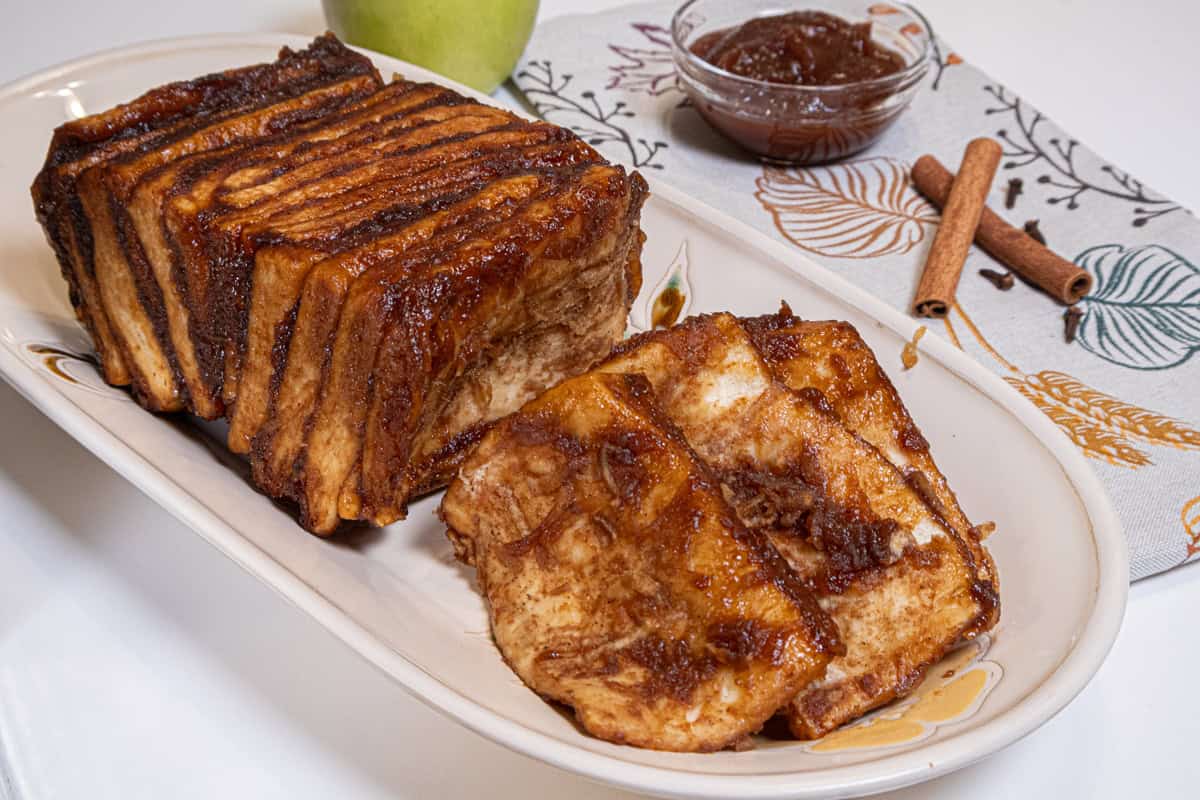 The Apple Cardamom Pull Apart Bread sits on a cream colored tray with an apple, apple butter, and cinnamon sticks in the background.