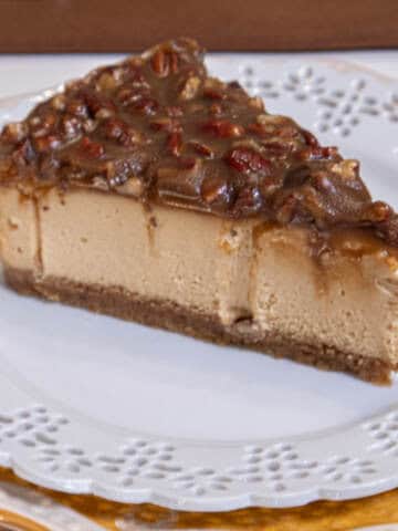 This Caramel Pecan Cheesecake sits on a white,laced edged plate on a gold and white plate with a brown napkin and fork in the background.
