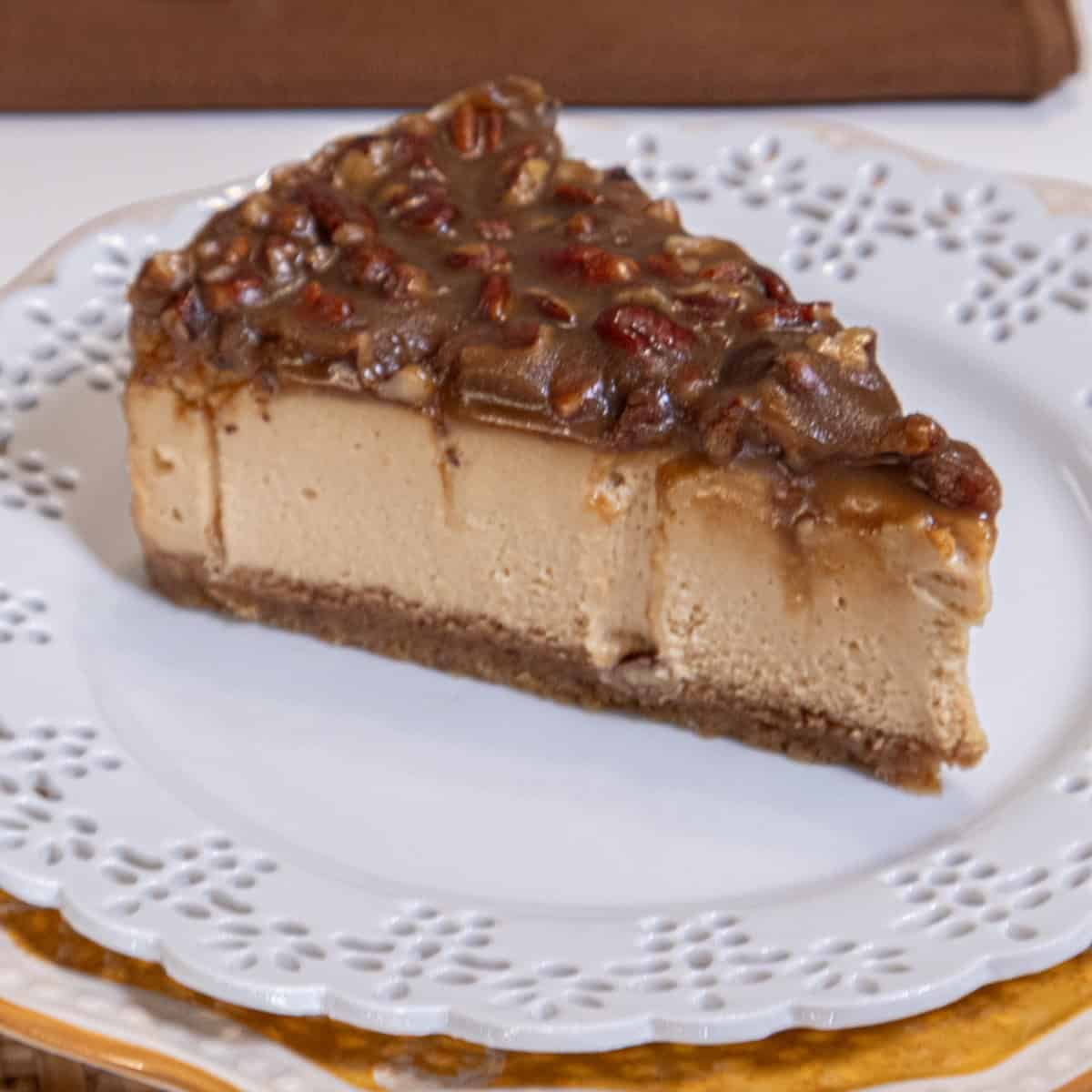 This Caramel Pecan Cheesecake sits on a white,laced edged plate on a gold and white plate with a brown napkin and fork in the background.
