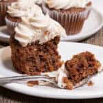 An Apple Spiced Cupcake with Maple Buttercream sits on a cream colored plate with a plate of more cupcakes behind it on a paisley cloth.