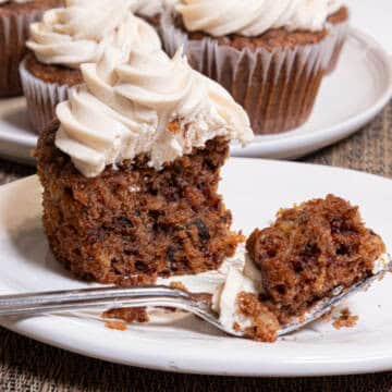 An Apple Spiced Cupcake with Maple Buttercream sits on a cream colored plate with a plate of more cupcakes behind it on a paisley cloth.