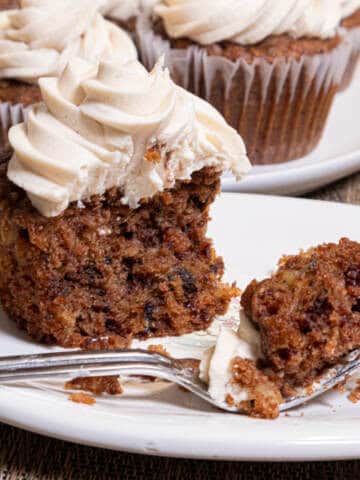 An Apple Spiced Cupcake with Maple Buttercream sits on a cream colored plate with a plate of more cupcakes behind it on a paisley cloth.