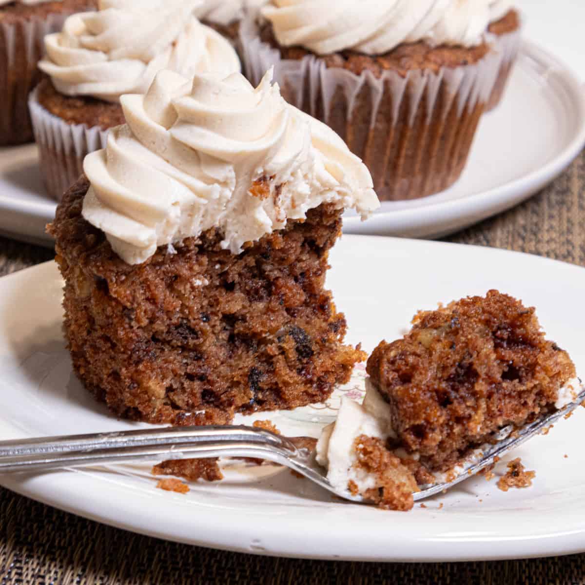 An Apple Spiced Cupcake with Maple Buttercream sits on a cream colored plate with a plate of more cupcakes behind it.
